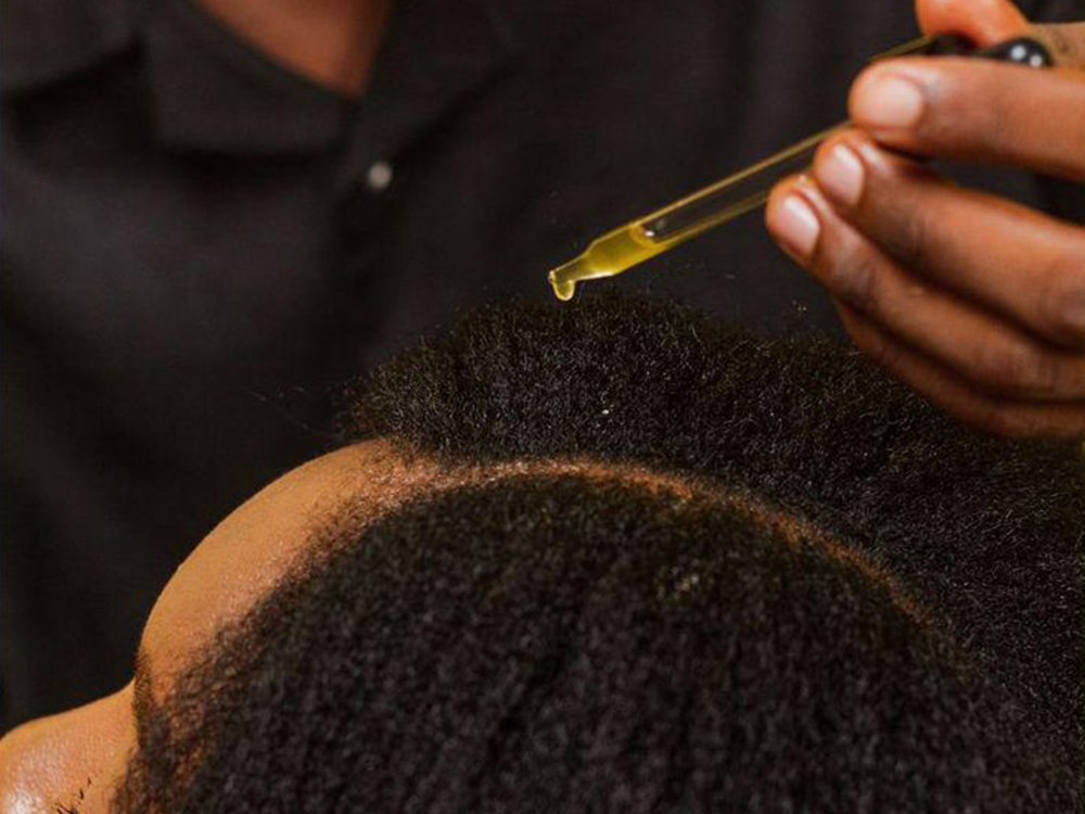 Bottle of JEJA Naturals Hair Growth & Hairline Repair Oil held above a grey-haired parting outdoors