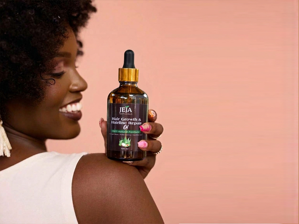Woman with a natural afro holding a bottle of JEJA Naturals Hair Growth & Hairline Repair Oil towards the camera